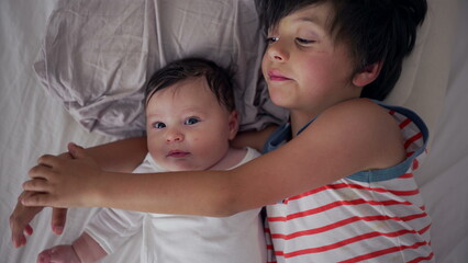 Older sibling lying on the bed with baby sibling, gently holding and bonding with them, creating a tender and intimate family moment
