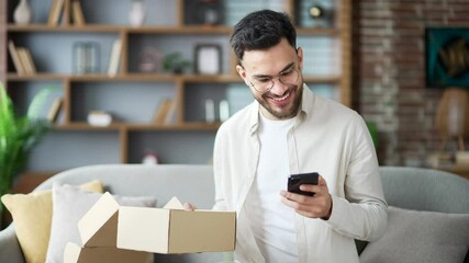 Joyful smiling young adult man customer opens package at home. Happy male writes good review on smartphone sitting on sofa in living room. Satisfied client of e-commerce. Express delivery services