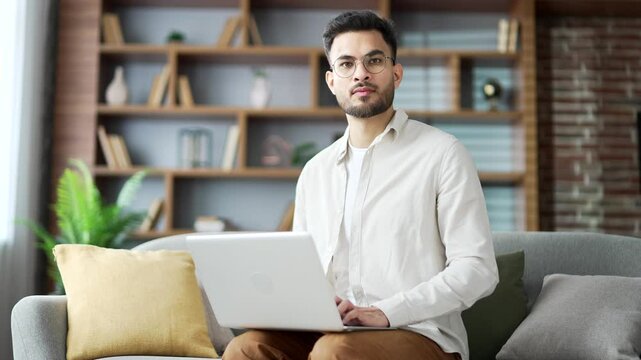 Handsome young adult man working typing on a laptop sitting on ofa in living room at home office. Bearded male freelancer using computer app, chatting online. busy with project looking at the camera