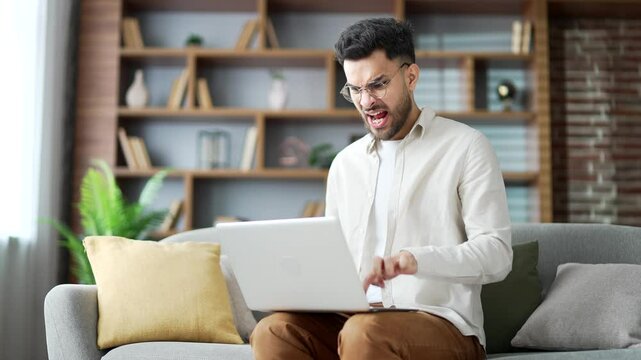 Angry frustrated young adult man having problems with software or service on laptop computer sitting on sofa in living room at home. Worried disappointed male complains about bad internet connection