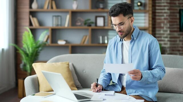 Happy handsome man dealing with household finances using website or app on laptop sitting at home office. Young adult male holds a utility bill and forms payments online by entering data on computer