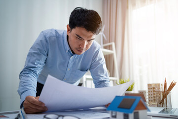 A man is sitting at a desk looking at a piece of paper. He is wearing a blue shirt and he is focused on the paper. The desk is cluttered with various items, including a potted plant, a cup