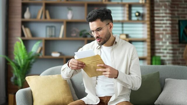 Disappointed handsome man reading letter with bad news sitting on sofa at home. Upset bearded male is depressed by an unpleasant notification, shocked by information in document looking at the camera