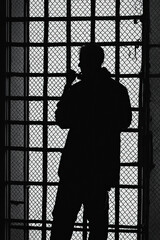 A striking silhouette of a man standing behind a mesh of prison bars, backlit by harsh light. The scene conveys themes of isolation, restriction, and emotional tension. Generative AI