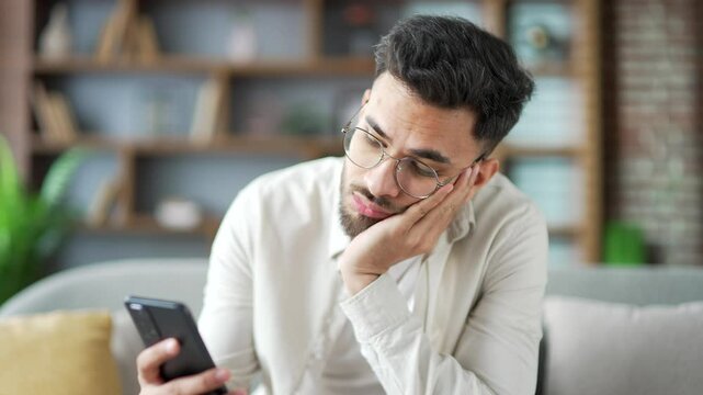 Lonely sad young adult male bored using smartphone sitting on sofa in living room at home. Tired sleepy handsome bearded man is depressed and in a bad mood, browses social media on a mobile phone