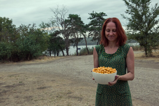 A young woman in green dress holds a bowl of yellow berries alycha in her hands.