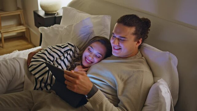 Laughing pair watching tablet movie cuddling in bed modern apartment closeup.