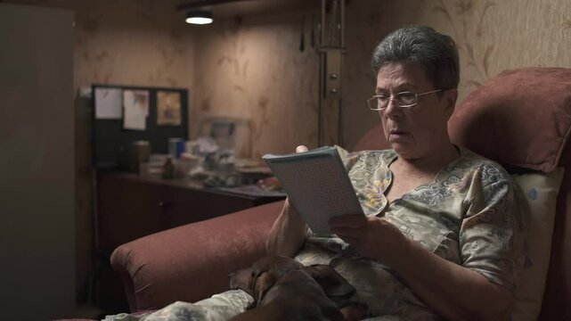 Elderly woman sitting on sofa and solving crossword puzzle, holding dachshund.