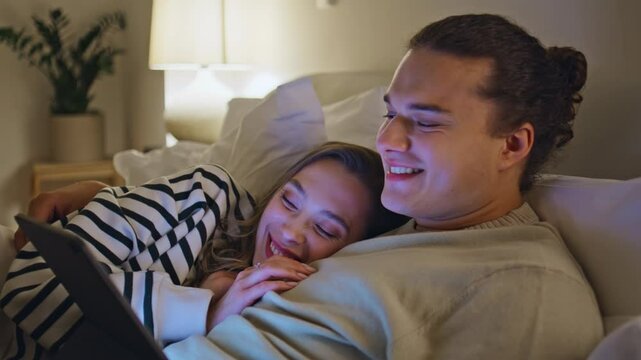 Joyful spouses looking tablet screen lying in bed enjoying time together closeup