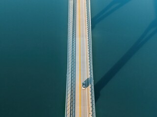Aerial view of a car driving across a bridge, casting long shadows over calm blue water, creating a minimalist scene of travel and solitude.