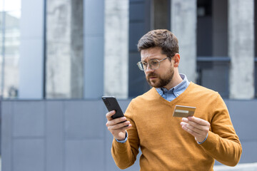 Close-up photo of a young man standing outside a building on the street, holding a credit card, looking upset and disappointed at a mobile phone screen