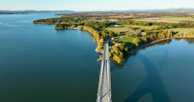 Aerial view of the Lake Champlain Bridge extending toward Vermont, with calm waters and a picturesque shoreline surrounded by autumn trees and rural farmland.