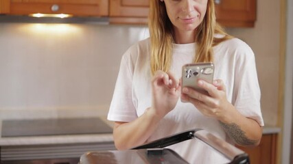 Woman using smartphone in kitchen while prepare meal. Stand at kitchen counter with sandwich maker and various ingredients laid out - Powered by Adobe