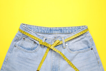 Jeans and measuring tape on yellow background, top view