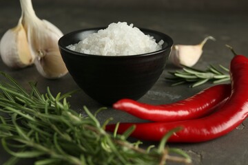 Sea salt in bowl, rosemary, chili peppers and garlic on grey table, closeup