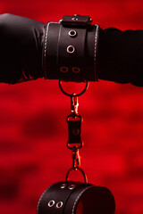 Close-up of female hands in black latex gloves and black leather BDSM handcuffs on a red...