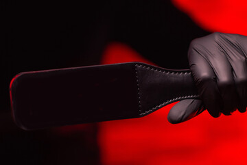 Close-up of female hands in black gloves holding a BDSM whip on a red background. BDSM is an outfit...