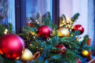 Festive window decor with red and gold Christmas ornaments and pine garland. Closeup of Xmas baubles and bulbs hanging on evergreen branches with twinkling lights.
