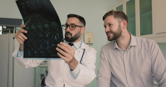 Male neurologist holding x-ray image explaining to patient diagnosis, provide consultation, share treatment options during his visit in clinic. Neurological disorders, injuries and diseases evaluation