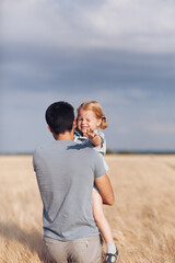 Young father carries his adorable ginger-haired little daughter in his arms through a wheat field