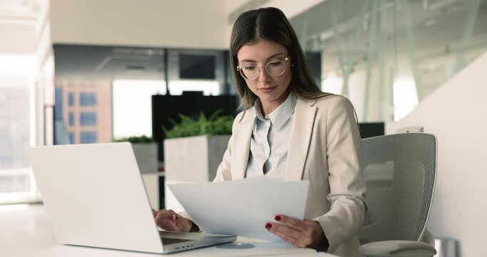 Focused business lady working in office seated at desk typing on laptop, check written information, holds documents, do paperwork, make report, busy in legal papers preparation for meeting or client