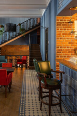 cozy vintage interior of the cafe restaurant with blue walls, wooden floors and brickwork. blue and red chairs are arrenged around tables. natural lighting and lamps. wrought-iron staircase leads up.