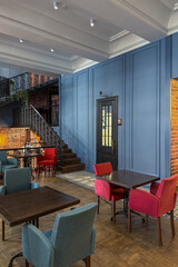 cozy vintage interior of the cafe restaurant with blue walls, wooden floors and brickwork. blue and red chairs are arrenged around tables. natural lighting and lamps. wrought-iron staircase leads up.