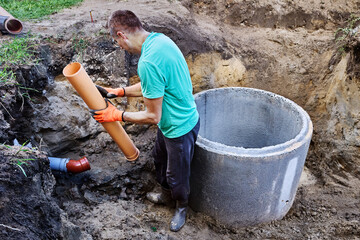 Connecting sewer pipe to an autonomous sewer system with septic tank made of concrete rings, worker takes measurements.