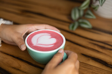 Young woman's hand holds a cup of matcha tea with raspberry or red berries, heart-shaped milk art, cappuccino coffee with red foam and heart, wooden table and decoration with plants, vegan hot drink