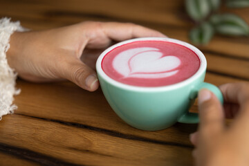 Young woman's hand holds a cup of matcha tea with raspberry or red berries, heart-shaped milk art, cappuccino coffee with red foam and heart, wooden table and decoration with plants, vegan hot drink