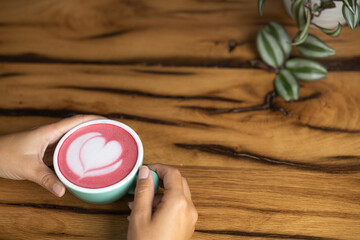 Young woman's hand holds a cup of matcha tea with raspberry or red berries, heart-shaped milk art, cappuccino coffee with red foam and heart, wooden table and decoration with plants, vegan hot drink