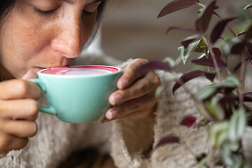 Close-up happy young woman smiling in a cafe holding a cup of matcha tea with raspberry, heart-shaped latte art, cappuccino coffee with red foam and heart, decoration with plants, vegan hot drink