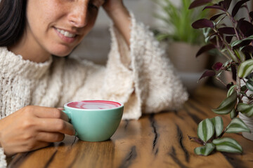 Close-up happy young woman smiling in a cafe holding a cup of matcha tea with raspberry, heart-shaped latte art, cappuccino coffee with red foam and heart, decoration with plants, vegan hot drink