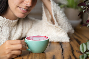 Close-up happy young woman smiling in a cafe holding a cup of matcha tea with raspberry, heart-shaped latte art, cappuccino coffee with red foam and heart, decoration with plants, vegan hot drink