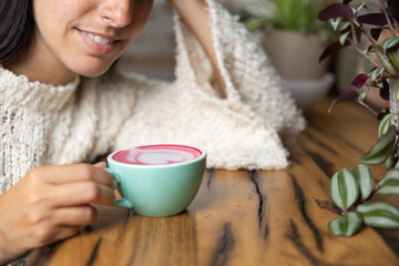 Close-up happy young woman smiling in a cafe holding a cup of matcha tea with raspberry, heart-shaped latte art, cappuccino coffee with red foam and heart, decoration with plants, vegan hot drink
