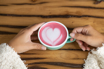 Young woman's hand holds a cup of matcha tea with raspberry or red berries, heart-shaped milk art, cappuccino coffee with red foam and heart, wooden table and decoration with plants, vegan hot drink