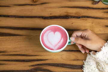 Young woman's hand holds a cup of matcha tea with raspberry or red berries, heart-shaped milk art, cappuccino coffee with red foam and heart, wooden table and decoration with plants, vegan hot drink