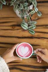 Young woman's hand holds a cup of matcha tea with raspberry or red berries, heart-shaped milk art, cappuccino coffee with red foam and heart, wooden table and decoration with plants, vegan hot drink