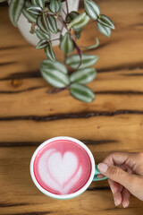 Young woman's hand holds a cup of matcha tea with raspberry or red berries, heart-shaped milk art, cappuccino coffee with red foam and heart, wooden table and decoration with plants, vegan hot drink