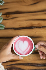 Young woman's hand holds a cup of matcha tea with raspberry or red berries, heart-shaped milk art, cappuccino coffee with red foam and heart, wooden table and decoration with plants, vegan hot drink