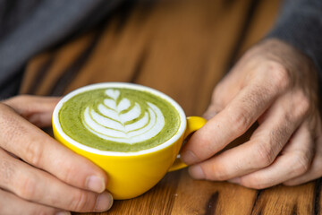 Young man hand holds a cup of matcha tea with raspberry or red berries, heart-shaped milk art, cappuccino coffee with red foam and heart, wooden table and decoration with plants, vegan hot drink