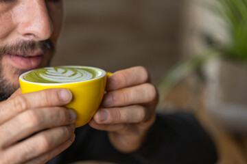 Close-up happy young man smiling in a cafe holding a cup of matcha tea with raspberry, heart-shaped latte art, cappuccino coffee with red foam and heart, decoration with plants, vegan hot drink
