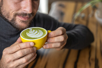 Close-up happy young man smiling in a cafe holding a cup of matcha tea with raspberry, heart-shaped latte art, cappuccino coffee with red foam and heart, decoration with plants, vegan hot drink