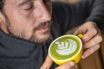 Close-up happy young man smiling in a cafe holding a cup of matcha tea with raspberry, heart-shaped latte art, cappuccino coffee with red foam and heart, decoration with plants, vegan hot drink