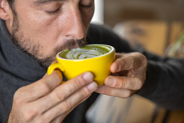 Close-up happy young man smiling in a cafe holding a cup of matcha tea with raspberry, heart-shaped latte art, cappuccino coffee with red foam and heart, decoration with plants, vegan hot drink