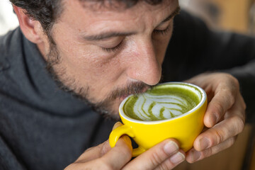 Close-up happy young man smiling in a cafe holding a cup of matcha tea with raspberry, heart-shaped latte art, cappuccino coffee with red foam and heart, decoration with plants, vegan hot drink