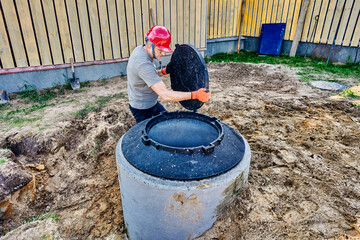New septic tank made of concrete rings is installed on house territory in rural area, worker carries manhole cover to cover well.