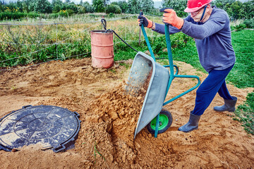 Worker unloads sand from wheelbarrow next to manhole cover of septic tank in rural area.