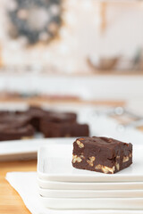 Piece of Homemade Chocolate Pecan Fudge on a White Plate with Tray of Fudge in Background in a Kitchen Decorated for Christmas