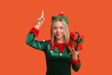Young woman in elf costume with Christmas gift box pointing at something on orange background